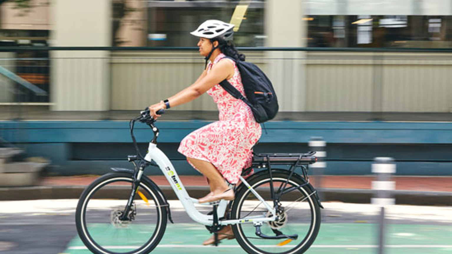 A woman in a pink floral dress rides an e-bike along a city street, the background is a blur.