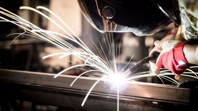 A person seen wearing a protective helmet who is welding a straight steel beam with a welding tool which is creating sparks where the tool and the steel are coming into contact.