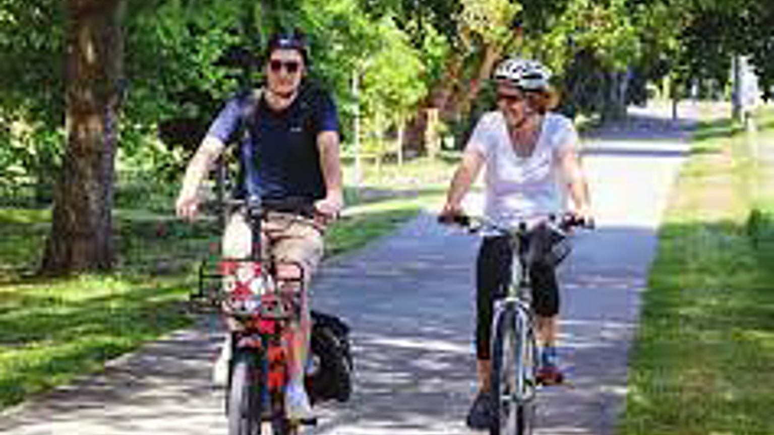 A man and a woman ride bikes along a flat, concrete path through a park.
